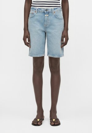 Άτομο που φοράει γαλάζια τζιν σορτς μέχρι το γόνατο, ριγέ τοπ και μαύρα σανδάλια με χρυσά διακοσμητικά σε σχήμα καρδιάς.