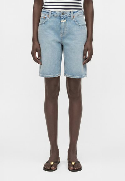 Personne portant un short en jean bleu clair jusqu'aux genoux, un haut rayé et des sandales noires avec des ornements dorés en forme de cœur.