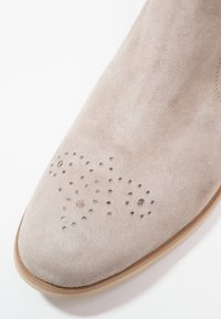 Bout de chaussure en daim beige clair avec perforations décoratives en motifs circulaires et en points, présentant un bord de semelle marron clair.