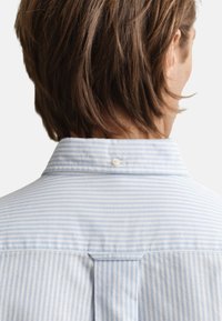 Mėlynos ir baltos spalvų dryžuota marškinėliai su sagomis ant apykaklės. Audinys atrodo minkštas, laisvo kirpimo, gale su klostės detalė.