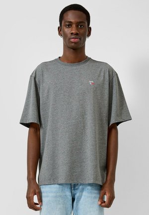 Junger Mann trägt ein locker sitzendes graues T-Shirt mit kleinem roten und weißen Text auf der Brust, kombiniert mit hellblauen Jeans, vor einem schlichten Hintergrund stehend.