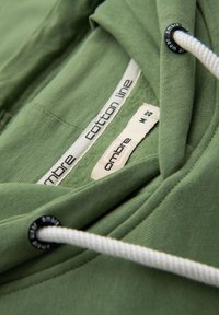 Sudadera verde de algodón con capucha y cordones blancos, etiqueta que dice "línea de algodón ombré", tamaño M y textura suave.