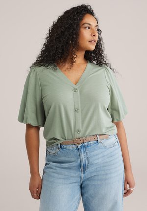 Frau mit lockigem Haar, die eine salbeigrüne Bluse mit Knöpfen und hellblaue, hoch taillierte Jeans mit einem beigen Gürtel trägt und zur Seite blickt.