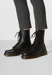 Botas de combate negras de cuero con cordones, acabado texturizado, puntera redonda y costuras amarillas sobre una suela gruesa y translúcida.
