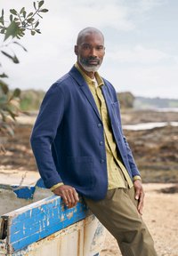 Marineblauer Baumwollblazer mit Knopfvorn und Taschen, über einem hellgrünen karierten Hemd getragen und mit olivgrünen Hosen kombiniert.