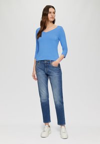 Blauwe top met driekwartmouwen gecombineerd met lichtblauwe, iets versleten jeans. Model draagt witte sneakers. Eenvoudige, casual outfit.