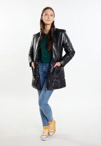 Giacca a vento nera con colletto alto, zip frontale e due tasche laterali, indossata sopra un cardigan verde, abbinata a jeans blue e sneakers gialle.