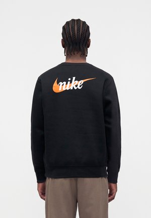 Fonott hajú férfi fekete pulóvert visel narancssárga Nike-logóval és fehér "nike" felirattal a hátán, egyszínű háttér előtt áll.