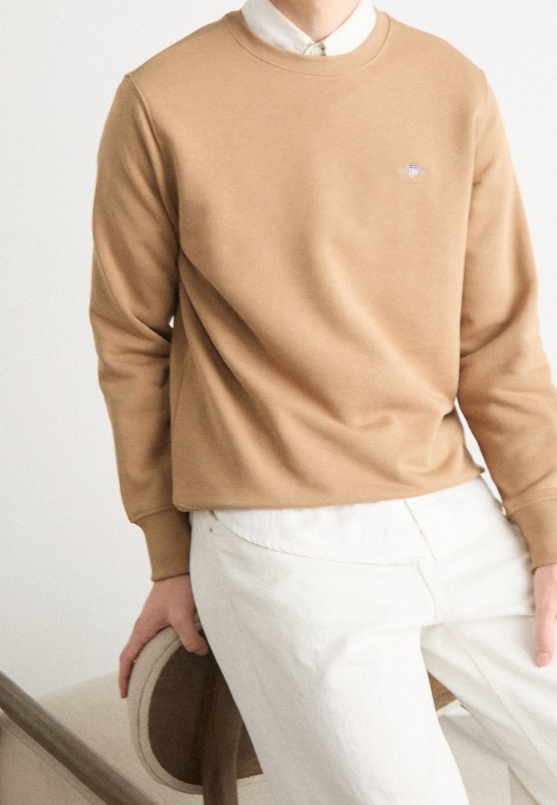 Personne portant un sweat-shirt beige par-dessus une chemise blanche à col et un pantalon blanc, assise sur une chaise beige avec une main posée dessus.