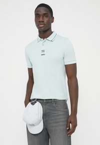 Φωτεινό γαλάζιο πόλο μπλουζάκι με μαύρες ρίγες στο γιακά και λογότυπο "BOSS", σε συνδυασμό με γκρι τζιν, κρατώντας ένα λευκό καπέλο με πράσινο έμβλημα.