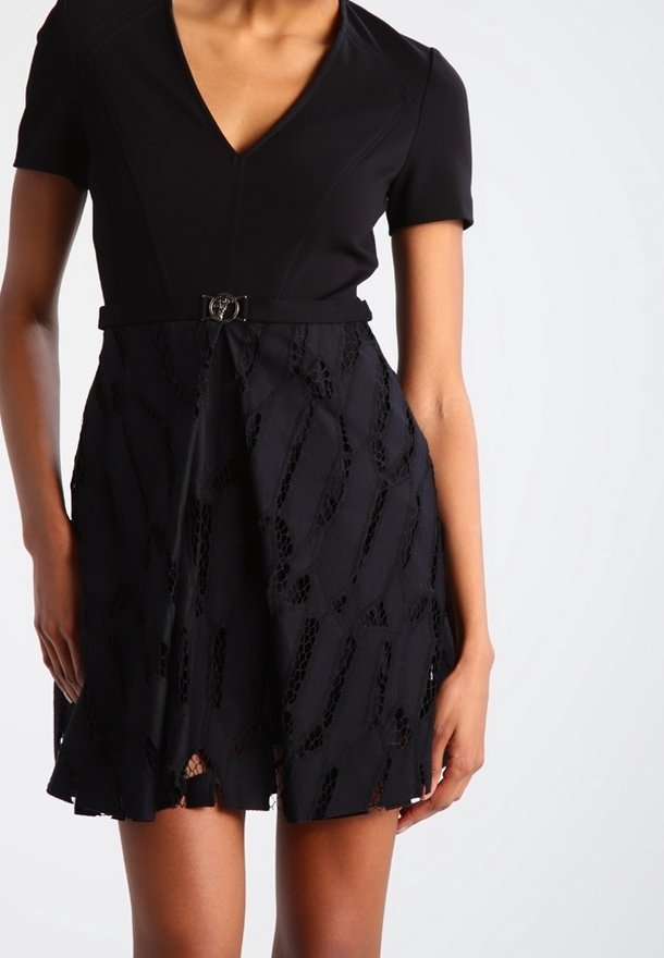 Femme portant une courte robe noire avec un col en V, des manches courtes et une jupe à motifs avec une petite ceinture à la taille.