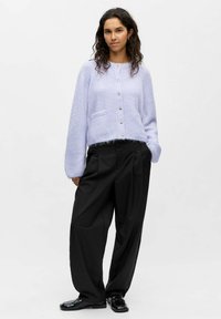 Jasnofioletowy, puszysty kardigan z przodu na guziki, z dwoma kieszeniami i luźnymi rękawami, w połączeniu z czarnymi szerokimi spodniami i błyszczącymi czarnymi butami.