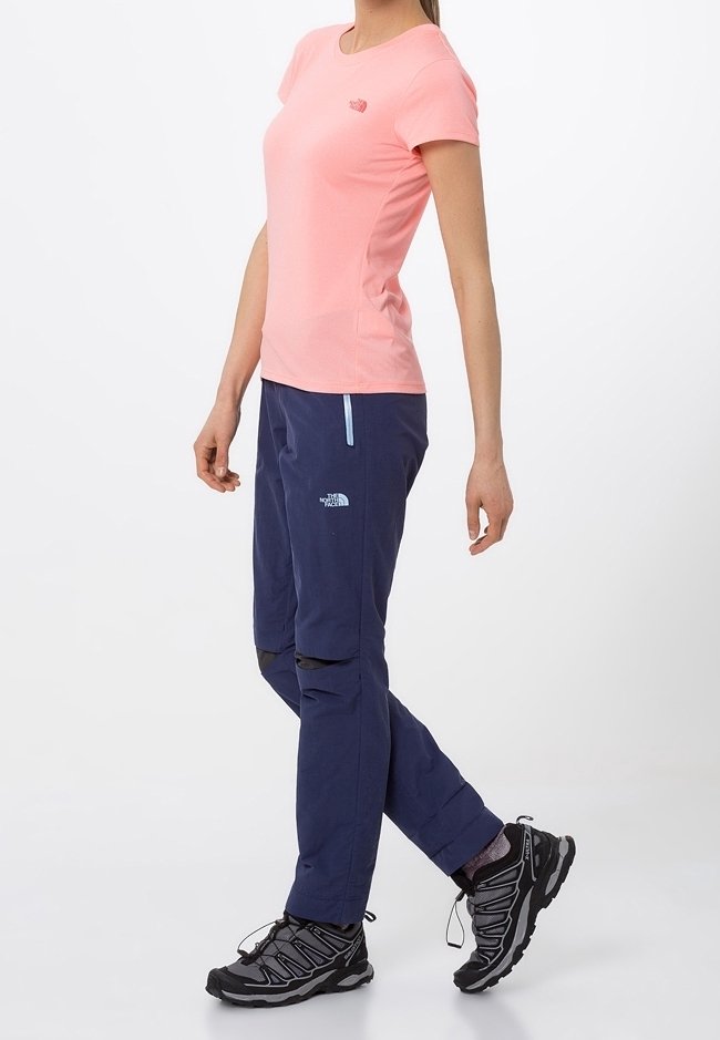 Light pink short-sleeve shirt with a small logo, paired with navy pants featuring zip pockets and a tapered design. Black hiking shoes with textured soles.