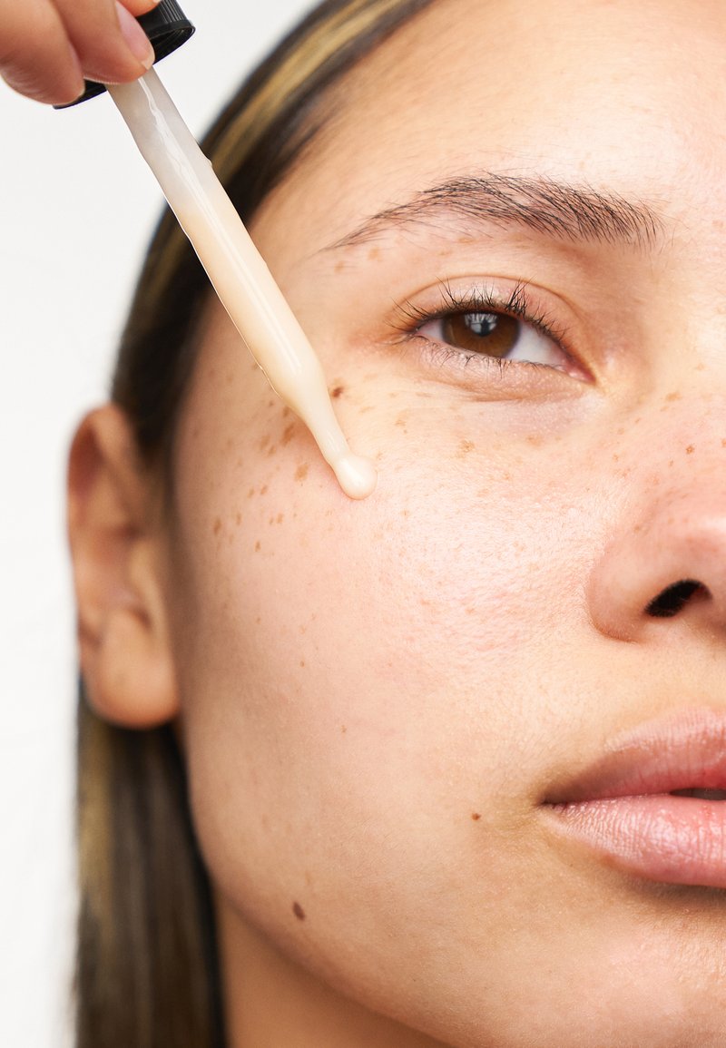 En dråpeflaske dispenserer en lys beige serum på huden med synlige fregner og jevn tekstur, og viser en klar hudtone og naturlig glød.