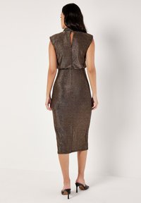 Vestido midi de bronce brillante con mangas cortas estructuradas, corte en forma de llave en la espalda y silueta ajustada, combinado con zapatos de tacón alto negros.