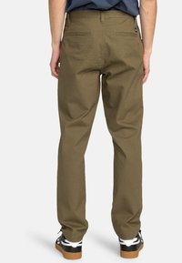 Khaki pants in sturdy cotton material, relaxed fit, with two back pockets and a waistband. Worn with black and white sneakers.
