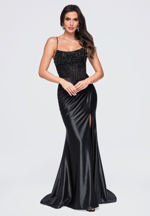 Femme portant une robe de soirée noire en satin ajustée avec un corsage orné de perles, des bretelles fines et une fente haute sur la cuisse, debout, les mains jointes devant elle.