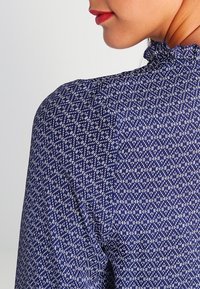 Navyblaue Bluse mit einem strukturierten Rautenmuster in Weiß; verfügt über einen rüschierten Kragen und ein tailliertes Design, das einen einfarbigen Rückansicht zeigt.