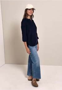 Marineblouse met driekwartmouwen, lichtblauwe flared jeans en bruine loafers. Model draagt een witte pet en staat tegen een neutrale achtergrond.