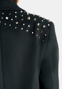 Cappotto di lana nero con borchie e gemme metalliche sulle spalle. Il tessuto ha una finitura morbida e strutturata.