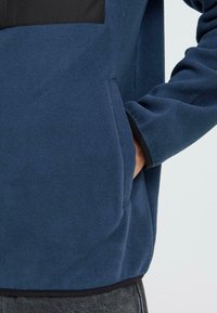 Chaqueta de forro polar navy con un bolsillo frontal negro, que presenta una textura suave y un dobladillo elástico. Incluye bolsillos laterales para mayor comodidad.