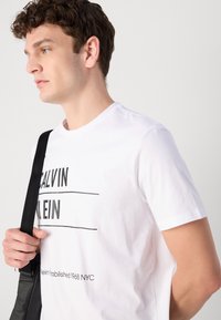 Bjela pamučna majica s crnim grafičkim tekstom i crtama. Kratki rukavi, okrugli izrez i opušteni kroj. Uparen s crnom torbom na remen.