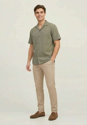 Uomo in piedi con le mani nelle tasche, indossa una camicia verde oliva a maniche corte, pantaloni beige e scarpe marroni, sorride verso la macchina fotografica.