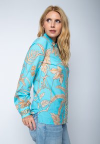 Turkusowa koszula z długim rękawem w beżowy kwiatowy wzór, z kołnierzykiem i zapięciem na guziki, w połączeniu z jasnoniebieskimi dżinsami.