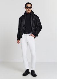 Schwarze Lederjacke mit Schaffellkragen, schwarzer Rollkragenpullover, weiße eng anliegende Hose und schwarze Chelsea-Stiefel. Dunkle Sonnenbrille vervollständigt den Look.