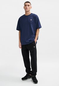 Navyblaue, übergroße T-Shirt mit kleinem Emblem, kombiniert mit schwarzen Trainingshosen, die ein ähnliches Emblem aufweisen, und schwarzen Sportschuhen.