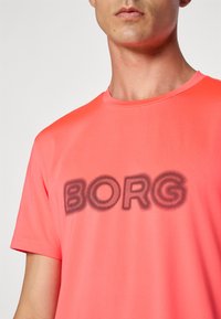 Mann som har på seg en lys korallfarget t-skjorte med svart prikket "BORG"-tekst sentrert på brystet mot en ensfarget bakgrunn.