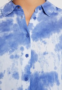 Chemise teinte bleu avec col, ornée de boutons assortis bleu, confectionnée dans un tissu doux au toucher lisse.