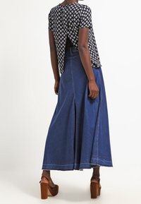 Top negro con estampado floral y una abertura en la parte posterior, combinado con una falda larga de mezclilla azul que presenta una abertura en la parte delantera, y sandalias de plataforma marrones.