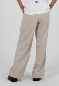 Personne portant un pantalon large beige et des chaussures noires, debout avec les mains derrière le dos devant un fond clair uni.