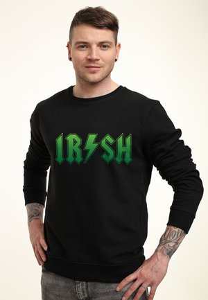 Młody mężczyzna z krótkimi brązowymi włosami, ubrany w czarną bluzę z zielonym napisem "IRISH" i grafiką pioruna, stoi z rękami na biodrach.