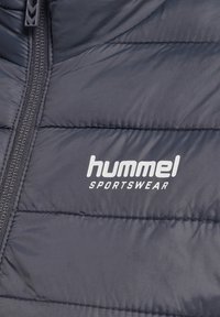 Marinblå isolerad jacka med sydda paneler, utrustad med dragkedja och en vit "hummel SPORTSWEAR"-logotyp på vänster sida.