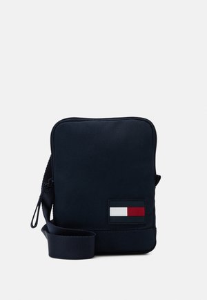 Kompaktní černá crossbody taška s nastavitelným pupkem a obdélníkovou červenobíločernou logo nášivkou na přední straně.