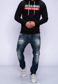 Felpa nera con logo bianco e rosso, abbinata a jeans blu strappati con lavaggio chiaro, completata da sneakers beige.