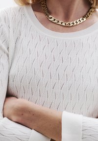 Witte, gebreide trui met een golfpatroon en een ronde halslijn. Gecombineerd met een glanzende, gouden ketting voor een accent.