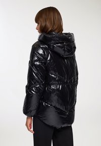 Giubbotto puffa nero con finitura lucida, dotato di cappuccio, polsini elasticizzati e dettagli di cucitura a zigzag. Materiale morbido e leggero.