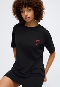 Μαύρη βαμβακερή μπλούζα t-shirt με κοντά μανίκια, χαλαρή εφαρμογή και κόκκινο εικονίδιο καρδιάς με το κείμενο "HUGO" στην αριστερή περιοχή του στήθους.