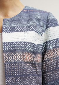 Veste tissée avec des rayures horizontales dans diverses nuances de bleu, blanc et corail. Tissu texturé avec des motifs complexes et un col arrondi.