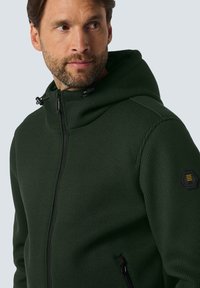 Groene zip-up hoodie van een gestructureerde stof, met een hoge kraag, verstelbare trekkoorden en een hexagonaal logo-patch op de mouw.