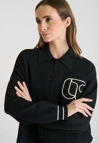 Cardigan nero con chiusura a bottoni, caratterizzato da un logo ricamato bianco sul petto e dettagli a righe chiare sui polsini.