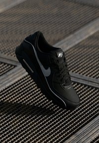 Sneaker Nike nera con tomaia testurizzata, lacci piatti, un logo prominente sulla linguetta e un accento grigio dello swoosh. Vista laterale su una superficie a griglia.