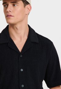 Camicia nera a maniche corte con un motivo testurizzato, dotata di colletto e cinque bottoni sul davanti, realizzata in un tessuto morbido.