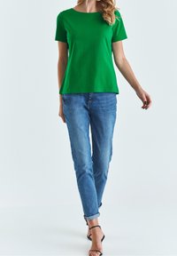 T-shirt verde a maniche corte in un tessuto liscio e leggero, con scollatura rotonda, abbinato a jeans blu chiaro a taglio affusolato e sandali neri.