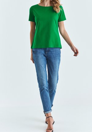 Grünes Kurzarm-T-Shirt aus einem glatten, leichten Stoff mit rundem Halsausschnitt, kombiniert mit hellblauen, tapered Jeans und schwarzen Sandalen.