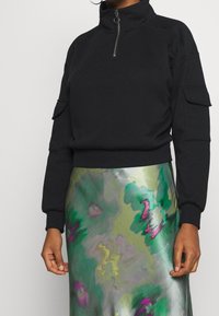 Svart pullover-tröja med hög krage, halv dragkedja och sidfickor. I kombination med en flerfärgad, glittrig kjol med gröna och lila mönster.