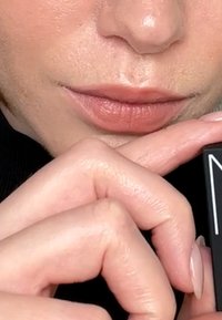 Ralentie nude avec une finition matte tenue entre des doigts. La main présente un teint de peau clair avec des ongles courts et naturels. Gros plan sur les lèvres.